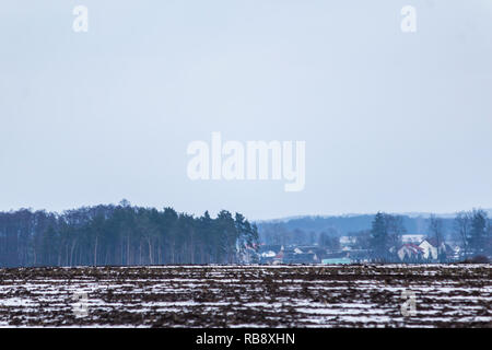 Un po' di neve sul campo arato. Villaggio e la foresta in background. L'inizio dell'inverno in Europa. Foto Stock