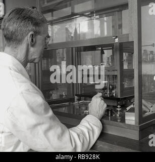 Degli anni Cinquanta, storico, un maschio maturo professore in un bianco-coat che lavora in un laboratorio di scienze dell'Università di Oxford, Inghilterra, facendo un esperimento utilizzando strumenti in una cornice di legno vetro protettivo armadio. Foto Stock