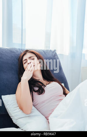 Giovane donna a sbadigliare e che copre la bocca con la mano sul letto durante la mattina tempo a casa Foto Stock