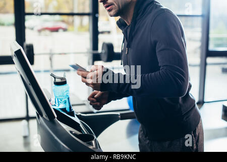 Immagine ritagliata di sportivo in hoodie esercita sul tapis roulant e utilizza lo smartphone in palestra Foto Stock