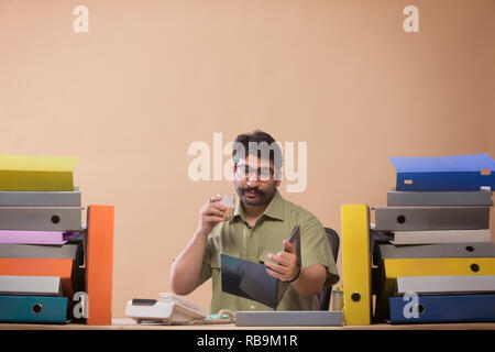 Imprenditore studiando i file mentre si bere il tè seduto alla sua scrivania in ufficio. Foto Stock