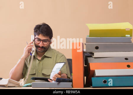 Imprenditore parlando telefonicamente mentre guardando i file seduto alla sua scrivania in ufficio. Foto Stock