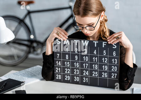 Bella imprenditrice vestiti di nero e occhiali seduti su una sedia e guardando il calendario Foto Stock