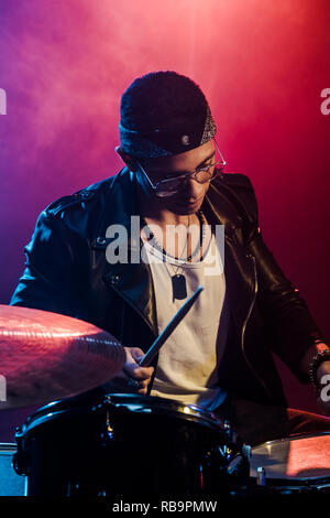 Grave giovane musicista maschio in giacca di pelle suonando la batteria durante un concerto rock sul palco con fumo e illuminazione drammatica Foto Stock