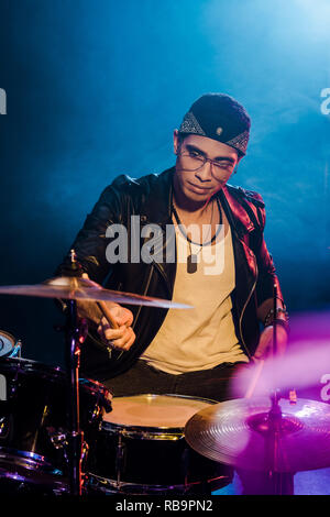 Fiducioso giovane musicista maschio in giacca di pelle suonando la batteria durante un concerto rock sul palco con fumo e illuminazione drammatica Foto Stock