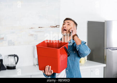 Uomo che parla sullo smartphone mentre si tiene secchio di plastica e guardando perde acqua dal soffitto Foto Stock