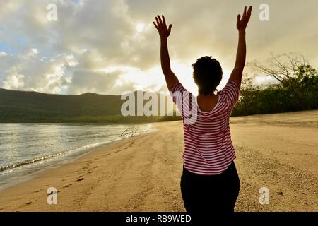 La donna trattiene le braccia in alto in libertà per celebrare sunrise su una spiaggia, spiaggia Smalleys Campeggio, Cape Hillsborough National Park, Qld, Australia Foto Stock
