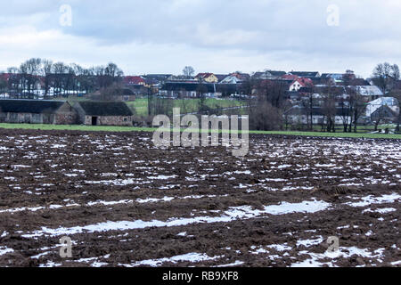 Un po' di neve sul campo arato. Villaggio e alberi in background. L'inizio dell'inverno in Europa. Foto Stock