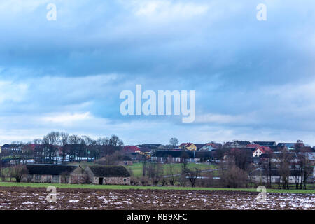 Un po' di neve sul campo arato. Villaggio e alberi in background. L'inizio dell'inverno in Europa. Foto Stock