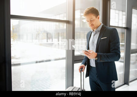 Bel giovane imprenditore stand in aeroporto hall. Egli tenere la mano della valigia e bianco tablet. Guy sorrisi. Egli stand in corrispondenza della finestra Foto Stock