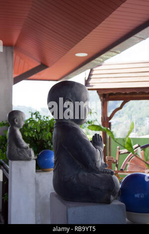 Due pregando buddha sculture Thailandesi recinzione terrazza con piante, palle blu e swing in legno sullo sfondo Foto Stock