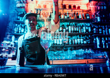 Bicchiere di fiery cocktail sul bancone del bar contro lo sfondo dei baristi mani con il fuoco. Barman giorno concept Foto Stock