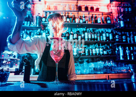 Bicchiere di fiery cocktail sul bancone del bar contro lo sfondo dei baristi mani con il fuoco. Barman giorno concept Foto Stock