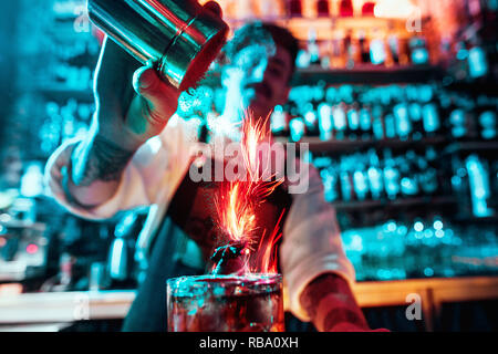 Bicchiere di fiery cocktail sul bancone del bar contro lo sfondo dei baristi mani con il fuoco. Barman giorno concept Foto Stock