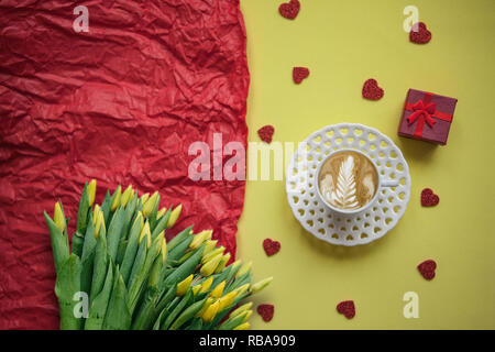 Un bicchiere di fresco aromatizzato caffè cappuccino con molti cuori e una scatola con un regalo. La prossima è un bouquet di tulipani gialli. Celebrazione del giorno di San Valentino o il giorno della donna o altro evento d'amore. Foto Stock