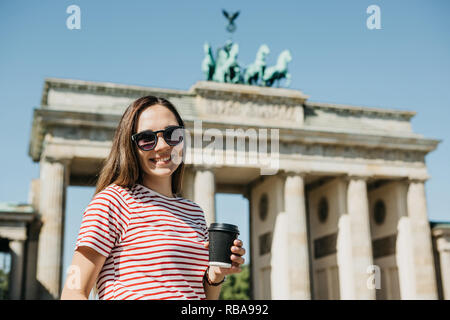 Ritratto di una giovane bella positivo ragazza sorridente che tiene in mano un caffè o altra bevanda sullo sfondo della Porta di Brandeburgo a Berlino in Germania. Foto Stock