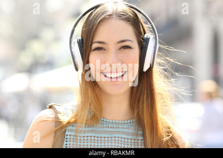 Vista frontale ritratto di una ragazza adolescente di indossare le cuffie per ascoltare musica guardando la telecamera in strada Foto Stock