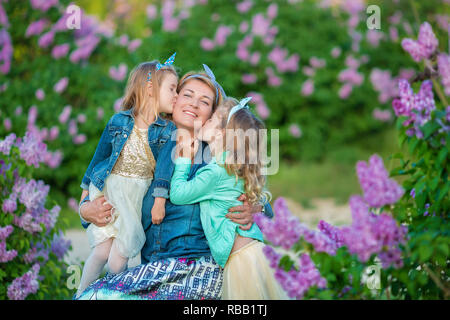 Madre donna con due cute ragazze sorridenti sorelle grazioso insieme su un campo di lilla bush tutti indossano abiti eleganti e jeans cappotti Foto Stock