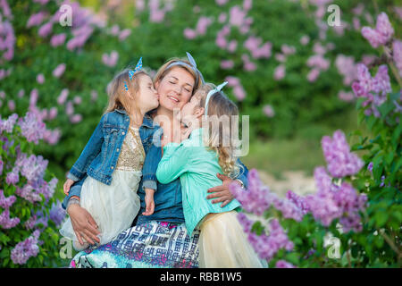 Madre donna con due cute ragazze sorridenti sorelle grazioso insieme su un campo di lilla bush tutti indossano abiti eleganti e jeans cappotti Foto Stock