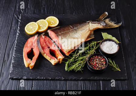 Il salmone selvatico tagliato in bistecche con rosmarino fresco sale, le foglie di alloro e grani di pepe su una pietra nera vassoio, vista da sopra Foto Stock