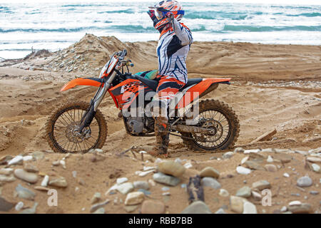 Vista posteriore del giovane uomo che guarda verso la fotocamera e seduto sul motociclo sulla spiaggia al tramonto Foto Stock