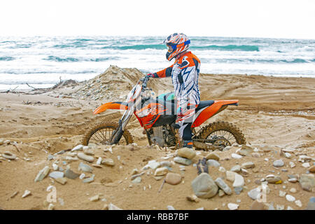 Vista posteriore del giovane uomo che guarda verso la fotocamera e seduto sul motociclo sulla spiaggia al tramonto Foto Stock