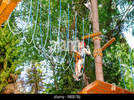 Brave elementari di età ragazza di indossare l'imbragatura di sicurezza arrampicate ad un corso di funi in outdoor parco avventura in una Foresta estate passando appendere la fune Foto Stock