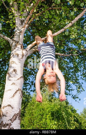 Basso angolo vista di felice ragazza appeso a testa in giù da un albero di betulla guardando la telecamera godendo di estate Foto Stock