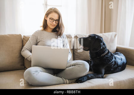 Ragazza con un cane seduto su un divano in interni, al lavoro su un notebook. Foto Stock