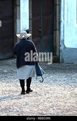 Un pomeriggio invernale in Vila Cova de Alva. Una signora anziana cammina giù per la collina dalla chiesa. Il umberella ombreggiatura era lei da sole d'inverno. Foto Stock