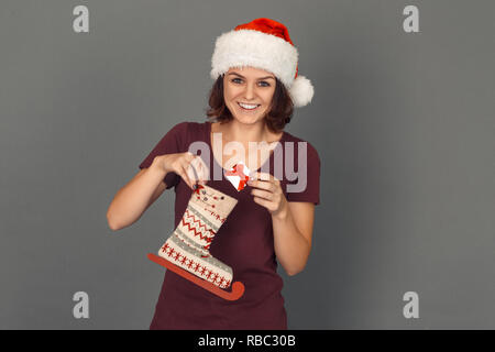 Giovane donna indossa santa hat tema natalizio isolato in piedi sul muro grigio messa piccola scatola regalo decorata in skate boot cerca fotocamera a sorridere c Foto Stock