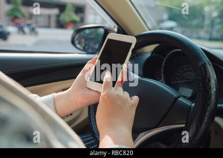 Femmina giovane conducente utilizzando il touch screen smartphone e mano che tiene il volante in un'auto. Foto Stock