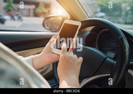 Femmina giovane conducente utilizzando il touch screen smartphone e mano che tiene il volante in un'auto. Foto Stock