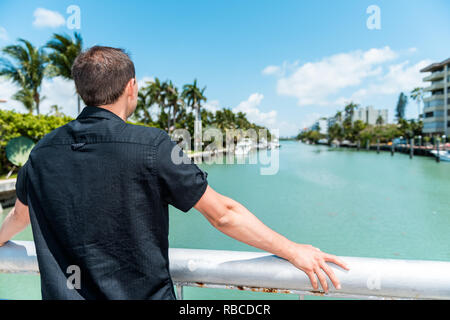 Giovane uomo torna indossando la maglia nera in piedi appoggiata sulla ringhiera del ponte in Bal Harbour, Miami Florida con oceano verde della Baia di Biscayne Foto Stock