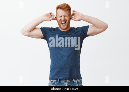 Orinato redhead guy sentire dolore in testa cause di rumore fastidioso suono, che copre le orecchie con le dita, gridando da dolorosa sensazione, chiudendo gli occhi e urlare con la bocca aperta oltre il muro grigio Foto Stock