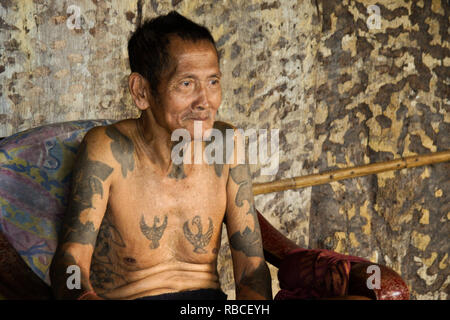 Anziani Iban uomo con tatuaggi Tribal seduto nella sedia in area comunale del Longhouse Mengkak, Batang Ai, Sarawak (Borneo), Malaysia Foto Stock