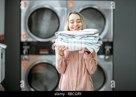 Giovane donna godendo pulire vestiti stirati in self serviced lavanderia con asciugatrice macchine sullo sfondo Foto Stock