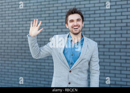 Ciao, è un piacere vedervi. Ritratto di felice bello uomo barbuto in stile casual in piedi e guardando la telecamera con il gesto di saluto e toothy sorridente. i Foto Stock