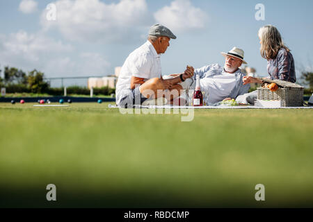 Gruppo di tre alti amici seduti in un parco godendo di cibo e bevande. Due alti uomini e una donna di mangiare snack durante una pausa al parco di bocce. Foto Stock
