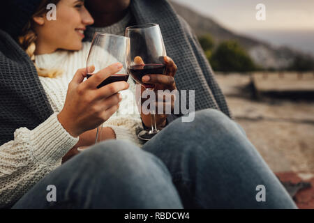 Ritagliato shot di un uomo e di una donna avvolta in una coperta seduti insieme vino di tostatura durante un picnic. Giovane godendo un picnic all'aperto con vino su un Foto Stock