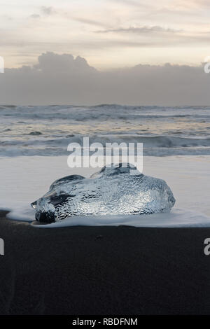 Round blocco di ghiaccio in sfumature di blu su una spiaggia con forte surf, al di sopra di esso uno strato debolmente cielo velato con colore giallo brillante tonalità e una formazione delle nuvole sulla hori Foto Stock