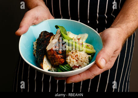 Close up di un chef mani presentando una ciotola di jerk pollo con riso e spezie e verdure Foto Stock