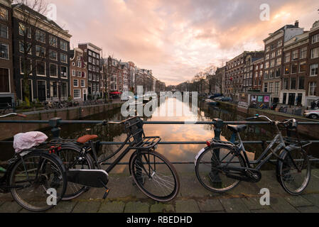 Biciclette bloccato alla barra di un ponte su un canale rivestito in architettura tradizionale case al tramonto nel centro di Amsterdam, Paesi Bassi. Foto Stock