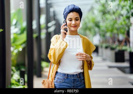 Una giovane e bella metà donna orientale in un turban hijab è sorridente come parla sul suo smartphone su strada durante il giorno. Foto Stock