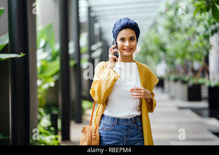 Una giovane e bella metà donna orientale in un turban hijab è sorridente come parla sul suo smartphone su strada durante il giorno. Foto Stock
