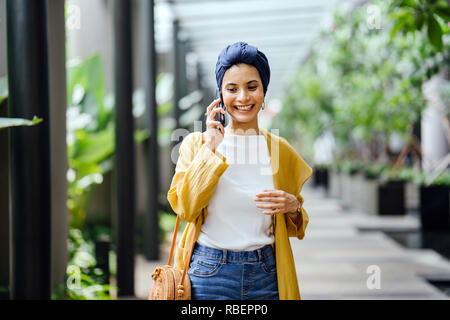 Una giovane e bella metà donna orientale in un turban hijab è sorridente come parla sul suo smartphone su strada durante il giorno. Foto Stock