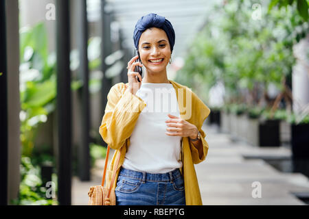 Una giovane e bella metà donna orientale in un turban hijab è sorridente come parla sul suo smartphone su strada durante il giorno. Foto Stock