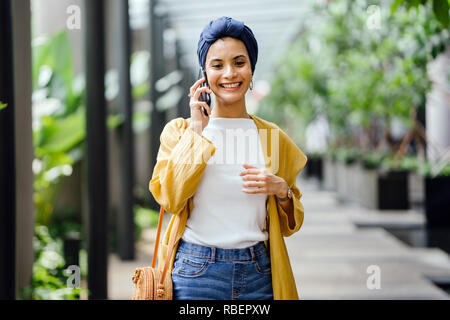 Una giovane e bella metà donna orientale in un turban hijab è sorridente come parla sul suo smartphone su strada durante il giorno. Foto Stock