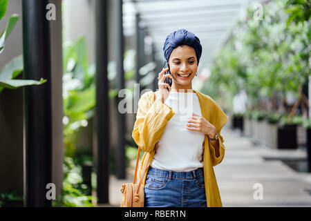 Una giovane e bella metà donna orientale in un turban hijab è sorridente come parla sul suo smartphone su strada durante il giorno. Foto Stock