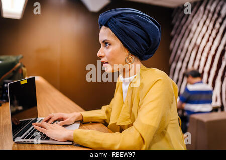 Un giovane malese donna asiatica è la digitazione e lavorare o studiare sul suo computer portatile in un caffè durante la giornata. Indossa un turbante hijab. Foto Stock